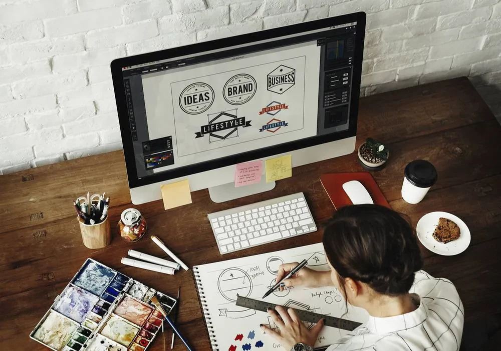A person sits at a wooden desk, sketching in a notebook with art supplies scattered around. A large computer monitor displays various logo designs labeled "Ideas," "Brand," and "Business." The workspace includes a cup of coffee, watercolor paints, and a tablet.