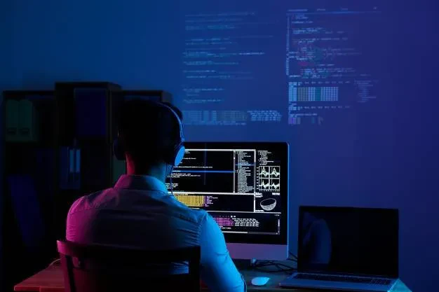 A person wearing headphones is sitting at a desk in a dimly lit room, working on a computer screen displaying multiple lines of code and data. A second monitor on the desk remains dark. The background shows projections of more code and data on the wall.