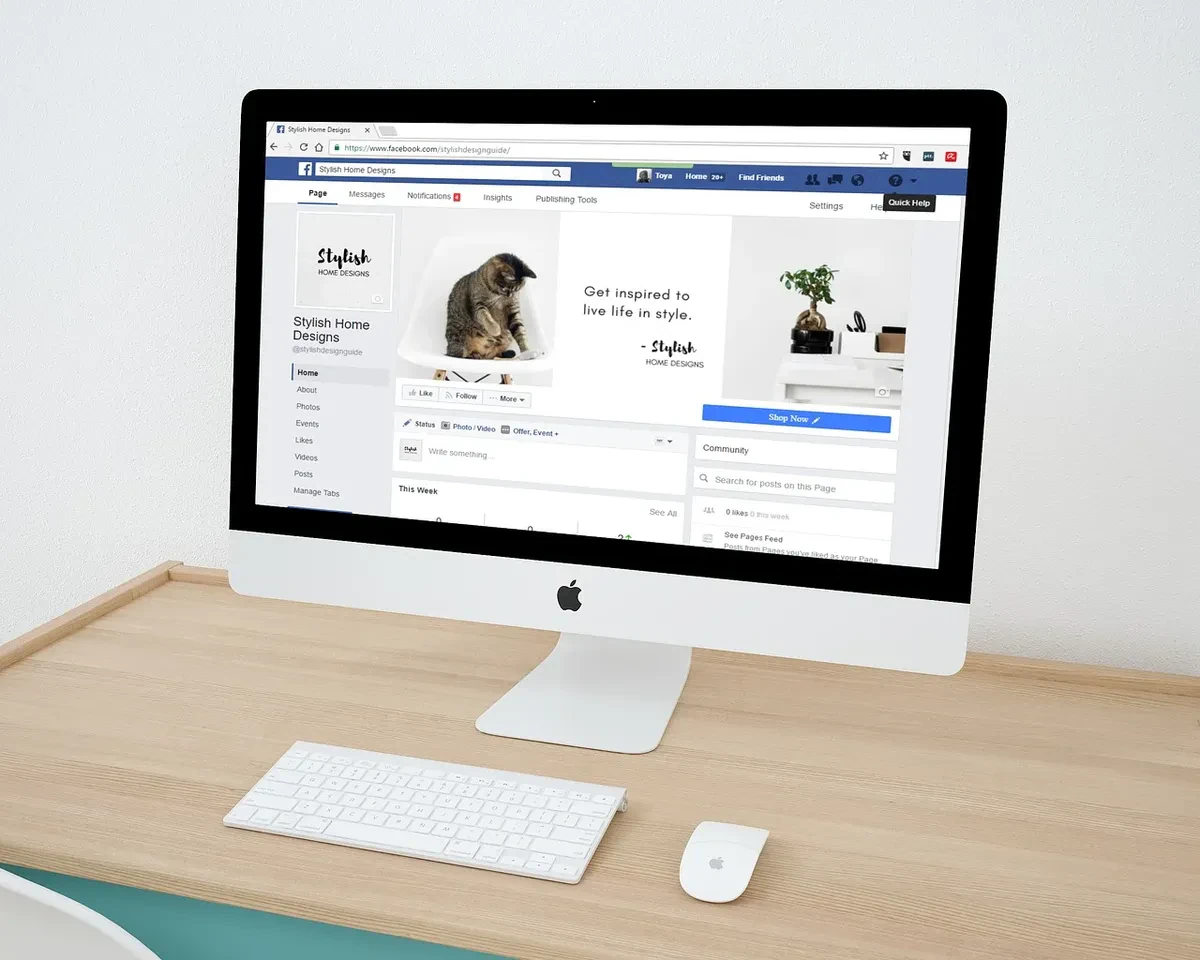 A sleek desktop computer with a keyboard and mouse is on a clean wooden desk. The screen displays a Facebook page titled "Stylish Home Designs" featuring a kitten and home decor items. The desk is minimalist with a white wall background.