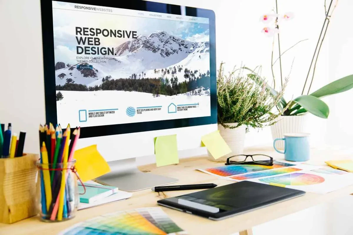 A well-organized workspace with a computer displaying a website about "Responsive Web Design." The desk has colorful pencils, sticky notes, glasses, a tablet, and color swatches. A potted plant adds a touch of greenery to the setup.