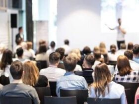 A large audience is seated in a conference room, facing a presenter who is speaking in front of a screen. The room is well-lit, and people appear to be attentively listening. The presenter is gesturing towards the screen, which displays a presentation.