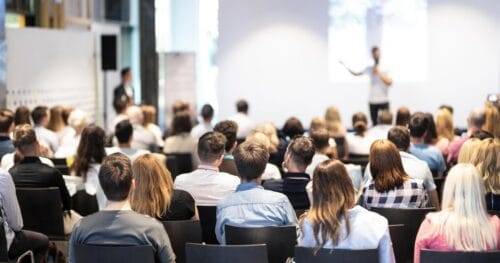 A large audience is seated in a conference room, facing a presenter who is speaking in front of a screen. The room is well-lit, and people appear to be attentively listening. The presenter is gesturing towards the screen, which displays a presentation.