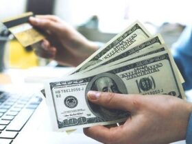 A person holding a stack of US dollar bills in one hand and a credit card in the other, sitting in front of a laptop computer. The scene suggests online shopping or financial transactions. The background includes a blurred-out office setting with office supplies.