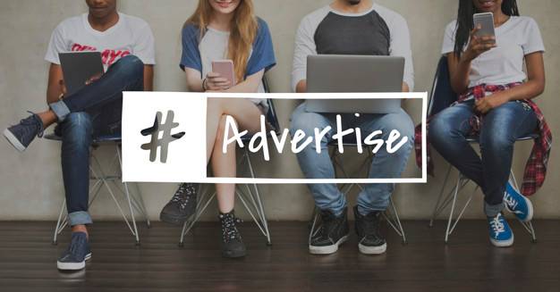Four young adults sit in a row, using various devices like smartphones and a laptop. An overlay text reads "#Advertise," implying a focus on digital media or marketing. They are casually dressed and seated against a plain wall with a wooden floor.