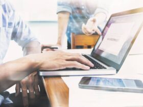 Close-up of two people at a desk. One is typing on a laptop, while the other is gesturing with their hand. A smartphone and papers are visible in the foreground. The scene is brightly lit, suggesting a work or study environment.