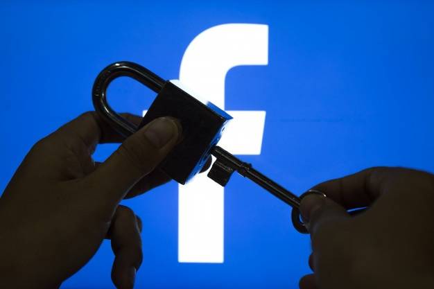 A pair of hands is holding a padlock and key in front of a large, illuminated Facebook logo. One hand is holding the padlock, while the other is inserting the key into the lock, suggesting the idea of unlocking or accessing Facebook.