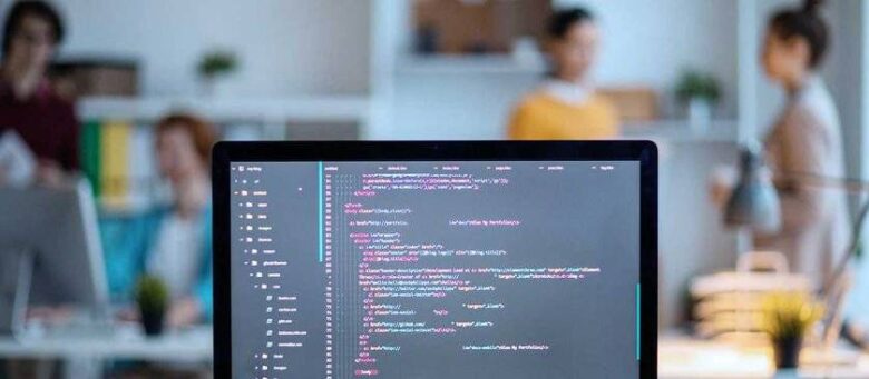 A computer screen displaying lines of code in a modern, brightly lit office setting. In the background, several people are engaged in conversation, with desks and office equipment visible. The focus is on the monitor, with the background slightly blurred.