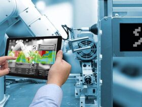 A person holds a tablet displaying a colorful data interface in front of an industrial robot arm. The robotic arm is part of a larger automated machine setup, with the focus on integrating digital and physical technologies in an industrial environment.