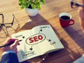 A person is sketching an SEO concept map on a notepad. The map includes terms like "Search," "Update," "Rankings," and "Link" connected to "SEO" in the center. On the table are eyeglasses, an open book, a potted plant, and a cup of coffee.