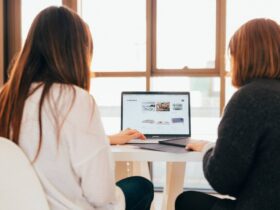 Two people are sitting at a table, facing a laptop. The screen shows a travel website with various travel-related images and options. They appear to be discussing or planning something, possibly a trip. The room is well-lit by large windows in the background.