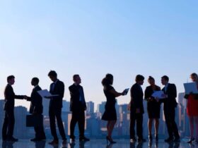 A diverse group of business professionals is standing in a line against a cityscape background. Some individuals are engaged in conversations, while others are examining documents or using electronic devices. The scene reflects a busy urban professional environment.