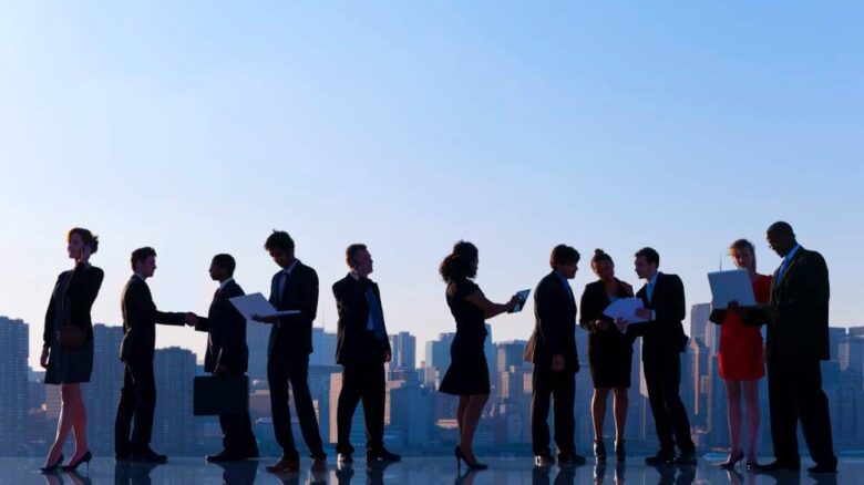 A diverse group of business professionals is standing in a line against a cityscape background. Some individuals are engaged in conversations, while others are examining documents or using electronic devices. The scene reflects a busy urban professional environment.