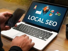 A person sits at a wooden table using a laptop displaying "LOCAL SEO" on the screen, with colorful icons representing digital marketing concepts. They hold a smartphone in their left hand and type on the laptop with their right. A black coffee cup is on the table.