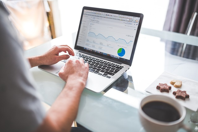 Person using a laptop displaying a graph and pie chart on the screen, indicative of data analysis or business analytics. There is a cup of coffee and a small plate with snacks next to the laptop on a glass-top table.