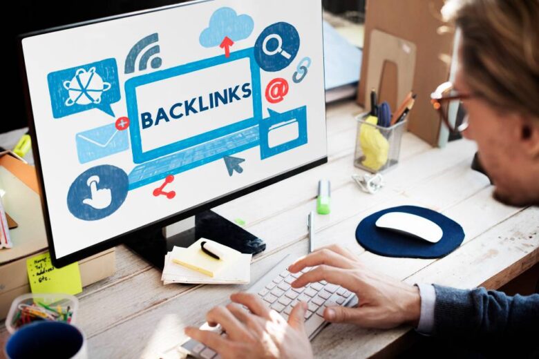 A person types on a keyboard at a desk with a computer screen displaying the word "BACKLINKS" surrounded by various icons like a cloud, email, link, and share symbol, indicating internet and digital communication themes. The workspace is bright and organized.