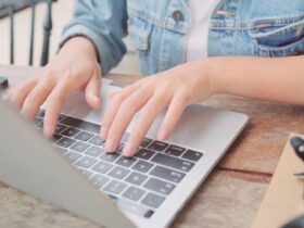Close-up of a person typing on a laptop keyboard. The person is wearing a denim jacket, and only their hands and forearms are visible. The setting appears to be a casual indoor environment, possibly a café, with a wooden table and a camera on the right side.