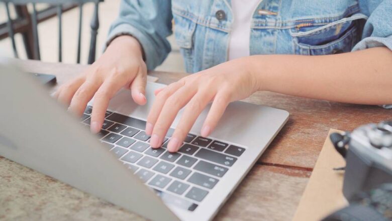 Close-up of a person typing on a laptop keyboard. The person is wearing a denim jacket, and only their hands and forearms are visible. The setting appears to be a casual indoor environment, possibly a café, with a wooden table and a camera on the right side.