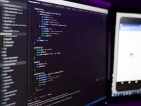 Two computer monitors are shown; the left screen displays code with syntax highlighting in an Integrated Development Environment (IDE), while the right screen shows a mobile application's interface. The workspace is dimly lit, highlighting the screens.