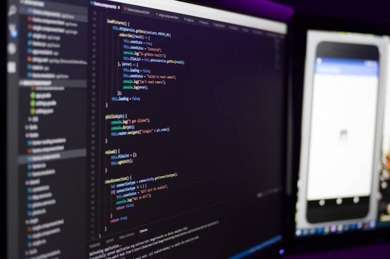Two computer monitors are shown; the left screen displays code with syntax highlighting in an Integrated Development Environment (IDE), while the right screen shows a mobile application's interface. The workspace is dimly lit, highlighting the screens.