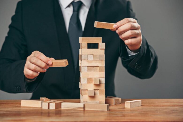 A person in a suit is building a tower from wooden blocks on a table. Their hands are placing a block on top of a partially constructed, unstable stack, with several blocks scattered around.