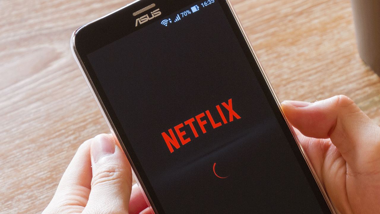 A person holds a smartphone displaying the Netflix app loading screen. The phone has an ASUS logo at the top and shows 70% battery and a time of 16:35. The iconic red Netflix logo is centered on the screen with a red loading circle below it.