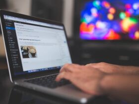 A person types on a laptop displaying a WordPress blog editor. The screen shows a blog post being created with a feature image of a coffee cup. The background is blurred, featuring a multicolored, illuminated screen.