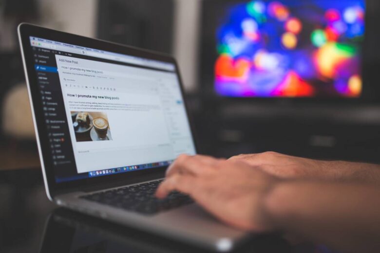 A person types on a laptop displaying a WordPress blog editor. The screen shows a blog post being created with a feature image of a coffee cup. The background is blurred, featuring a multicolored, illuminated screen.