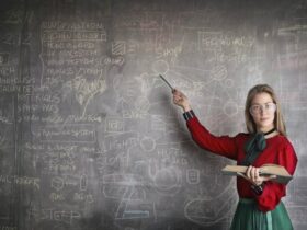 A person with long hair and glasses, wearing a red and green outfit, stands in front of a chalkboard covered in various diagrams and notes. They hold an open book in one hand and point to the chalkboard with a pointer in the other hand.