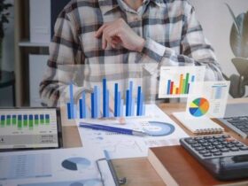 A person wearing a checkered shirt sits at a desk analyzing various charts and graphs on papers and electronic devices. A tablet, notebook, calculator, and other documents are on the desk. Holographic bar and pie charts are visibly displayed above the papers.