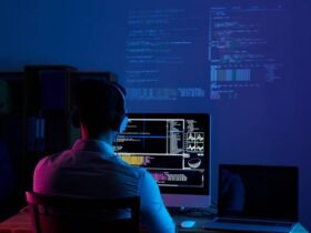 A person wearing headphones is sitting at a desk in a dimly lit room, working on a computer screen displaying multiple lines of code and data. A second monitor on the desk remains dark. The background shows projections of more code and data on the wall.