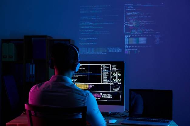 A person wearing headphones is sitting at a desk in a dimly lit room, working on a computer screen displaying multiple lines of code and data. A second monitor on the desk remains dark. The background shows projections of more code and data on the wall.