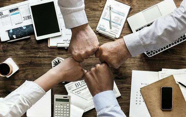 Four individuals fist bump over a desk filled with work items, including a tablet, smartphone, newspapers, notebook, and calculator, symbolizing teamwork and collaboration.