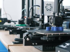 A row of 3D printers on a wooden desk, each in the process of printing an object. The focus is on the nearest printer producing a blue cylindrical shape. The background shows additional 3D printers and related equipment in a well-lit room.