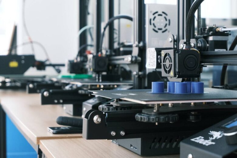 A row of 3D printers on a wooden desk, each in the process of printing an object. The focus is on the nearest printer producing a blue cylindrical shape. The background shows additional 3D printers and related equipment in a well-lit room.