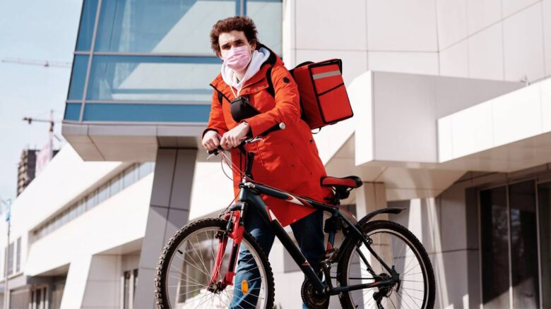 A person wearing a red coat and a pink face mask is standing next to a black bicycle. They have a red delivery bag on their back and are in an urban area with modern buildings in the background.
