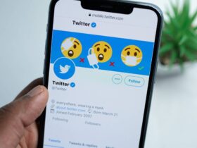 A hand holds a smartphone displaying a Twitter profile. The profile's header image shows emojis with and without masks, emphasizing mask-wearing. The blue Twitter logo is visible, and the profile mentions wearing a mask, with a join date of March 2007.