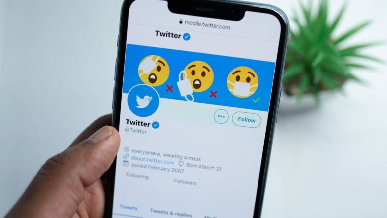 A hand holds a smartphone displaying a Twitter profile. The profile's header image shows emojis with and without masks, emphasizing mask-wearing. The blue Twitter logo is visible, and the profile mentions wearing a mask, with a join date of March 2007.
