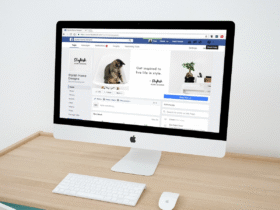 A sleek desktop computer with a keyboard and mouse is on a clean wooden desk. The screen displays a Facebook page titled "Stylish Home Designs" featuring a kitten and home decor items. The desk is minimalist with a white wall background.
