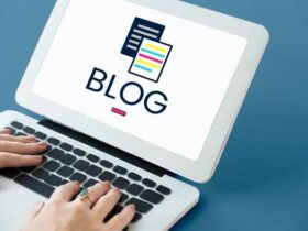 A person types on a laptop displaying the word "BLOG" in bold letters on the screen, accompanied by an icon of two overlapping documents. The background is a plain blue surface.