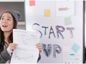 A woman wearing a knit hat holds papers with charts and graphs, standing in front of a whiteboard that has "START UP" written on it. The whiteboard also has colorful sticky notes and printed charts attached to it.