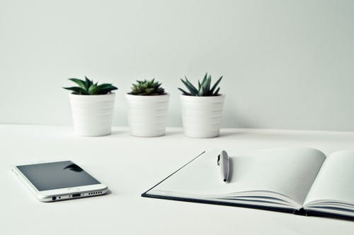 A white desk with a smartphone, an open notebook with a pen placed on top, and three small potted succulents arranged in a row against a light-colored wall. The arrangement appears neat and minimalist.