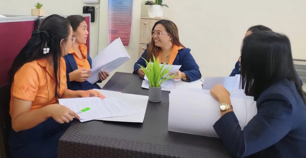 Group of women in orange and navy uniforms sitting around a table in a workplace, discussing documents during a team meeting.