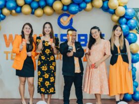 A group of five people stands on a stage in front of a festive balloon arch made of blue and gold balloons. They are dressed in coordinated outfits featuring orange, black, and yellow tones. The person in the center holds a trophy, while others hold red envelopes. Behind them is a backdrop with the logo and text "WINNERS" partially visible, suggesting an award or recognition event. All are smiling and posing for the photo.