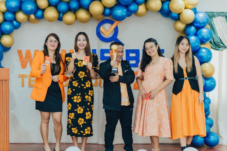 A group of five people stands on a stage in front of a festive balloon arch made of blue and gold balloons. They are dressed in coordinated outfits featuring orange, black, and yellow tones. The person in the center holds a trophy, while others hold red envelopes. Behind them is a backdrop with the logo and text "WINNERS" partially visible, suggesting an award or recognition event. All are smiling and posing for the photo.